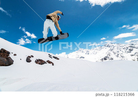 snowboarder jumps from a big air with a board grab, a nose or tail trick in rotation, against the backdrop of snow-capped mountains and the bright sun 118215366