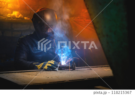 A welder in a leather jacket and welding mask welds in a dark room of an old barn. Home welding 118215367