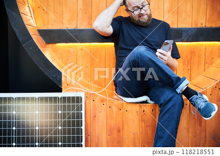 Bearded man using smartphone connected to photovoltaic solar panel. Integration of sustainable renewable energy into everyday life, showing practical use of solar power. Cropped view. Focus on phone. 118218551