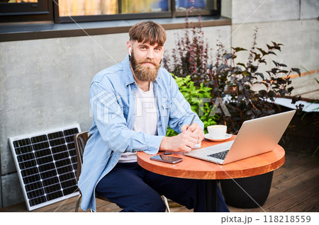 Portrait of bearded man works at round wooden table with laptop, notebook, and cup of coffee. Guy wears earbuds, surrounded by greenery and solar panel, highlighting modern, productive workspace. 118218559