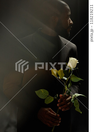 Vertical shot of African American gentleman in elegant suit holding white rose sadly looking away while grieving loss of loved one on black background in studio, ghost glare effect Vertical shot of African American gentleman in elegant suit holding white rose sadly looking away while grieving loss of loved one on black background in studio, ghost glare effect 118219211