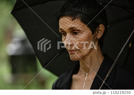 Portrait of grieving senior woman in mourning veil looking down at gravestone while hiding from rain under black umbrella, copy space Portrait of grieving senior woman in mourning veil looking down at gravestone while hiding from rain under black umbrella, copy space 118219250