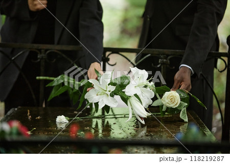 Cropped shot of unrecognizable people placing white lilies on memorial stone slab wet from rain to commemorate deceased person at cemetery, copy space 118219287