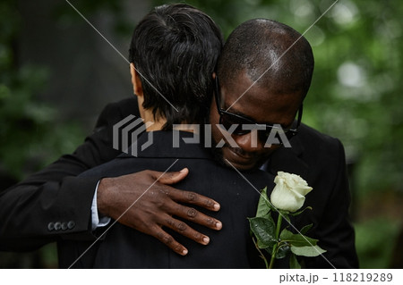 Medium shot of adult Black man in sunglasses hugging senior woman while grieving over loss of loved one at cemetery Medium shot of adult Black man in sunglasses hugging senior woman while grieving over loss of loved one at cemetery 118219289