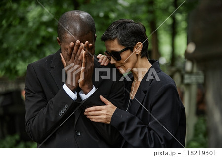 Medium shot of senior woman in sunglasses consoling crying husband at memorial ceremony. Multiethnic couple grieving loss of loved one at cemetery 118219301