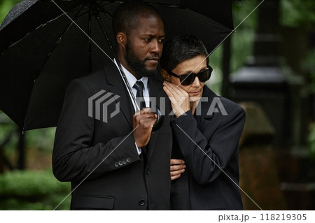 Medium shot of mourning multiethnic couple grieving at memorial ceremony. Senior woman clinging to Black mans shoulder hiding from rain under black umbrella at cemetery 118219305
