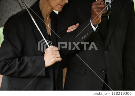 Cropped shot of senior woman in black suit holding husband by arm while hiding from rain under umbrella, copy space 118219306