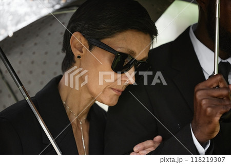 Close up of grieving senior woman in black sunglasses standing by husband holding supportive man by arm, while hiding from rain under umbrella at memorial service at cemetery Close up of grieving senior woman in black sunglasses standing by husband holding supportive man by arm, while hiding from rain under umbrella at memorial service at cemetery 118219307