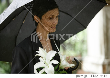 Side view of senior woman under black umbrella wearing mourning veil and grieving loss of departed loved one at grave while holding white lilies at cemetery, copy space 118219316