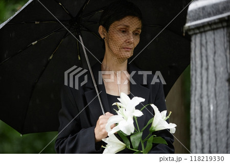 Medium shot of senior woman in mourning veil holding white lilies visiting grave of passed away loved one, while hiding from rain under umbrella at cemetery Medium shot of senior woman in mourning veil holding white lilies visiting grave of passed away loved one, while hiding from rain under umbrella at cemetery 118219330