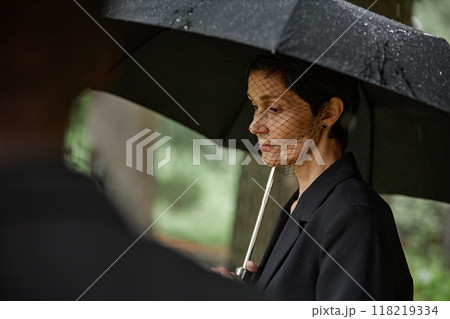 Side view of senior woman in black suit and veil grieving over loss of loved one at memorial ceremony hiding from rain under umbrella at graveyard, copy space Side view of senior woman in black suit and veil grieving over loss of loved one at memorial ceremony hiding from rain under umbrella at graveyard, copy space 118219334