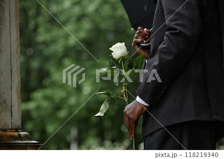 Cropped shot of Black man in funeral black suit holding white rose in memory of deceased loved one before placing flower on grave in rainy cemetery, copy space Cropped shot of Black man in funeral black suit holding white rose in memory of deceased loved one before placing flower on grave in rainy cemetery, copy space 118219340