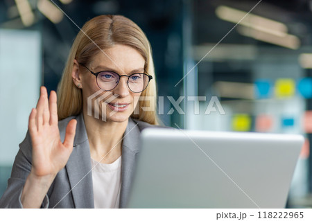 Professional woman in office using laptop for video call. She waves hand at screen, engaged in virtual communication. Glasses and suit reflect work environment. Technology enhances teleconference. 118222965