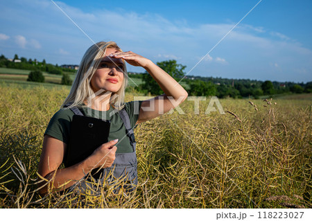 a woman farmer standing in the middle of rapeseed field, observing the horizon, shielding herself from the sun with hand and holding a tablet 118223027