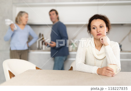 Frustrated woman sitting in kitchen during quarrel between her husband and elderly mother. 118227835