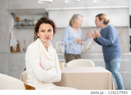 Sad thinking woman sits in kitchen and listens to man quarrel with elderly woman 118227993