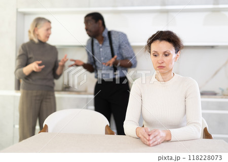 Middle-aged woman sitting at the kitchen table with her back to man and old woman quarreling together Middle-aged woman sitting at the kitchen table with her back to man and old woman quarreling together 118228073