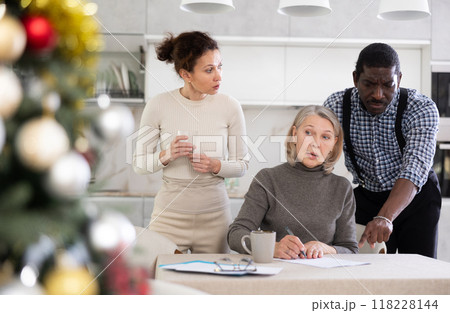 Old woman compiling testament while middle-aged man and woman competing with each other in the kitchen with Xmas-tree Old woman compiling testament while middle-aged man and woman competing with each other in the kitchen with Xmas-tree 118228144