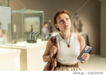Positive woman visitor with smartphone in hands exploring religious art in museum 118228145