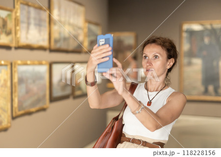 Interested woman looking closely at paintings in art gallery, using smartphone and taking photo 118228156