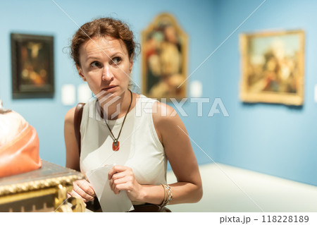 Brunette woman visitor holding guide list standing near pictures in museum of arts Brunette woman visitor holding guide list standing near pictures in museum of arts 118228189