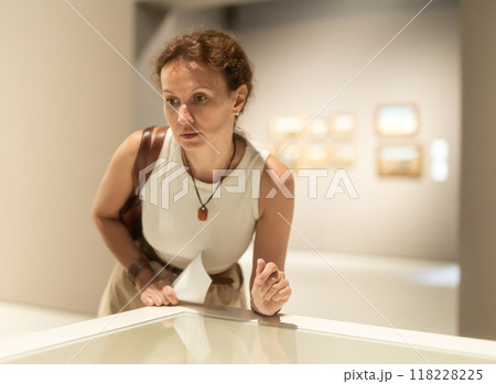 Attentive woman standing near exposition and looking with interest at art objects under glass in museum 118228225