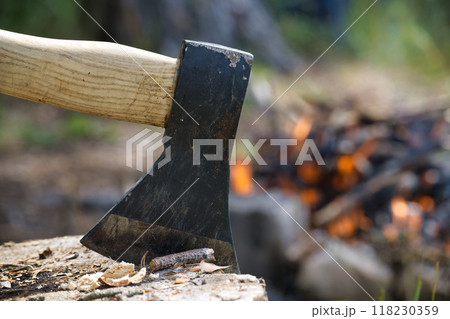 Close-up of an axe embedded in a log with a campfire burning in the background 118230359