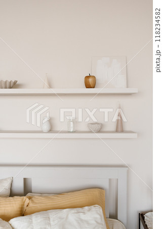 White bed with headboard and a yellow pillow, surrounded by shelves with various items on them, including vase, bowl and apple 118234582