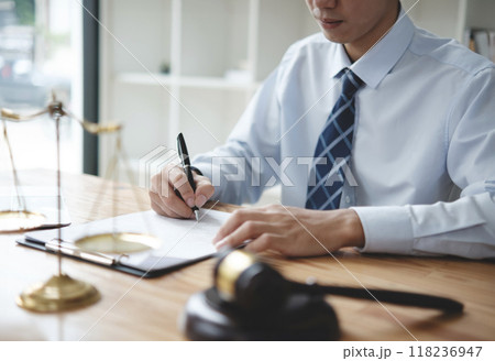 A lawyer in a suit is sitting at his desk, signing legal documents with a gavel on the side. 118236947