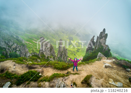 Aerial view of hiker stands triumphantly with arms raised on misty mountain ridge, surrounded by dramatic rock formations. Fog partially obscures view, adding mystical atmosphere to scene. Aerial view of hiker stands triumphantly with arms raised on misty mountain ridge, surrounded by dramatic rock formations. Fog partially obscures view, adding mystical atmosphere to scene. 118240216