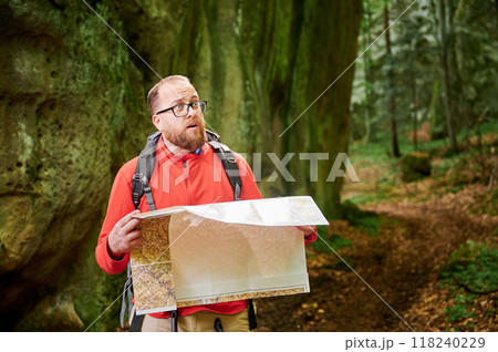 Bearded tourist man with surprised expression studies map while stands in front of large rock in dense forest. Traveler with grey backpack and glasses, planning outdoor adventure, hiking route. Bearded tourist man with surprised expression studies map while stands in front of large rock in dense forest. Traveler with grey backpack and glasses, planning outdoor adventure, hiking route. 118240229