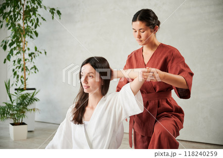 Masseuse making thai yoga massage. Woman therapist doing traditional massage treatment for female patient indoors. Client sitting, meditating, enjoying hand massage in calm atmosphere. 118240259