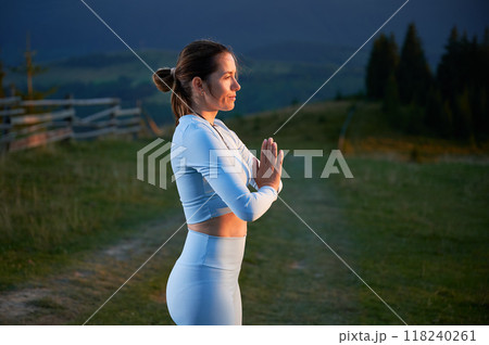 Woman practicing yoga and meditating outdoors in the mountains in a serene, natural setting. Female performing yoga pose, with backdrop of beautiful mountain landscape at sunrise or sunset. 118240261