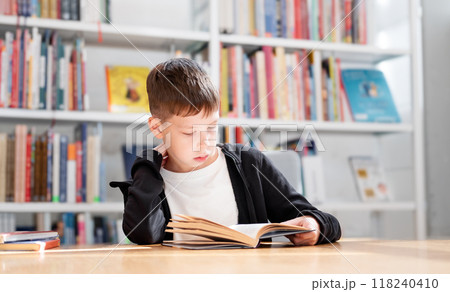 11-year-old teenage boy in a white T-shirt reads an interesting book in the library against the background of bookshelves 11-year-old teenage boy in a white T-shirt reads an interesting book in the library against the background of bookshelves 118240410