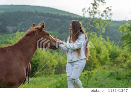 A woman is petting a brown horse in a grassy field. The scene is peaceful and serene, with the woman and the horse sharing a moment of connection. 118241347