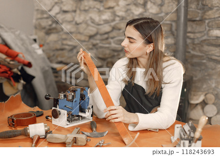 Young woman working in her workspace. Woman wearing an apron and making a belt. Grounge dark stone texture background. Young woman working in her workspace. Woman wearing an apron and making a belt. Grounge dark stone texture background. 118243693