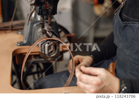 Cropped photo of mature craftsman working in his workspace. Man wearing an apron and using a sewing machine. Grounge dark stone texture background. 118243706