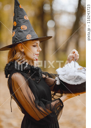 Blonde young girl witch in forest on Halloween. Girl wearing black dress and cone hat. Witch holding a magician stuff. Blonde young girl witch in forest on Halloween. Girl wearing black dress and cone hat. Witch holding a magician stuff. 118243723