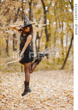 Brunette young girl witch in forest on Halloween. Girl wearing black dress and cone hat. Witch holding a magician stuff. 118243724