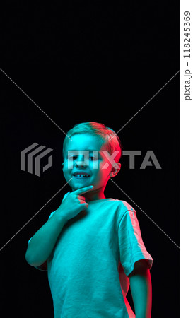 Vertical portrait of cheerful smiling boy, child in t-shirt posing with thoughtful positive face against black studio background in neon light Vertical portrait of cheerful smiling boy, child in t-shirt posing with thoughtful positive face against black studio background in neon light 118245369