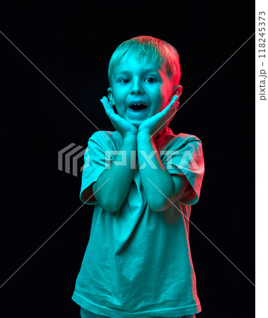 Portrait of cute little boy, child in casual white t-shirt holding hand to cheeks and smiling against black studio background in neon light 118245373