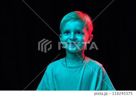 Portrait of little 4 years old boy with blonde hair, standing with cheerful smile, wearing t-shirt and posing against black studio background in neon light 118245375