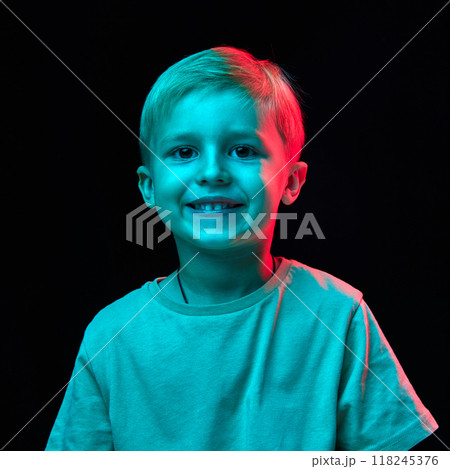 Portrait of little 4 years old boy with blonde hair, standing with cheerful smile, wearing t-shirt and posing against black studio background in neon light Portrait of little 4 years old boy with blonde hair, standing with cheerful smile, wearing t-shirt and posing against black studio background in neon light 118245376