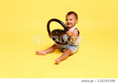 Beautiful little baby, girl sitting in a denim outfit, holding steering wheel, smiling with pure joy against yellow studio background Beautiful little baby, girl sitting in a denim outfit, holding steering wheel, smiling with pure joy against yellow studio background 118245399