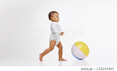 Little baby, girl in onesie walking confidently towards colorful ball, playing with curiosity and joy isolated on white studio background Little baby, girl in onesie walking confidently towards colorful ball, playing with curiosity and joy isolated on white studio background 118245401