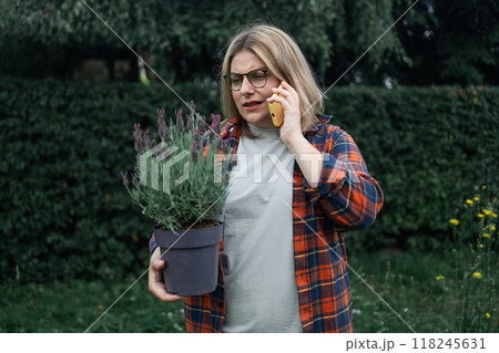 Shot of an happy female Caucasian farmer talking on the smartphone and holding a plastic pot of lavender flower plants to plant in the ground. Gardener using mobile phone 118245631