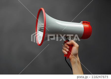 A close-up image of a person's hand holding a red and white megaphone against a dark background, symbolizing communication, announcement, and amplification 118247873
