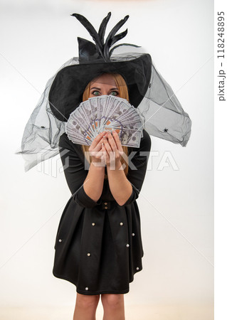 Woman is posing wearing a witch costume for Halloween, holding a stack of us dollar bills in front of her face Woman is posing wearing a witch costume for Halloween, holding a stack of us dollar bills in front of her face 118248895