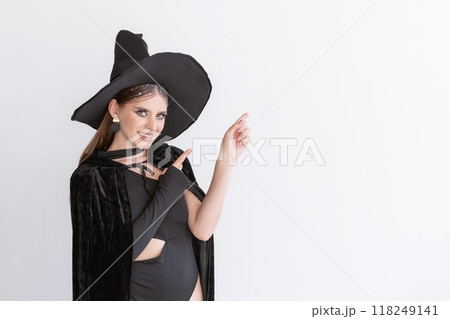 Beautiful smiling woman in black hat and cloak points her index finger up. White background. Space for text. Black Friday. Halloween party Beautiful smiling woman in black hat and cloak points her index finger up. White background. Space for text. Black Friday. Halloween party 118249141