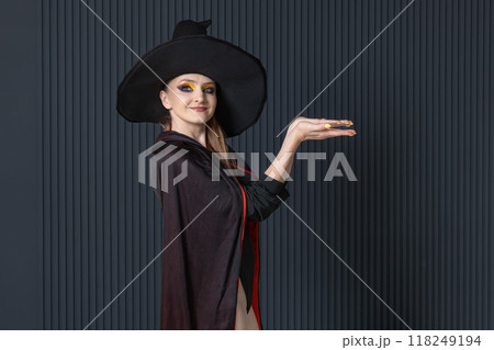 Beautiful young woman in a witch hat shows something against a dark wall. Halloween party, costume evening. Place for text 118249194